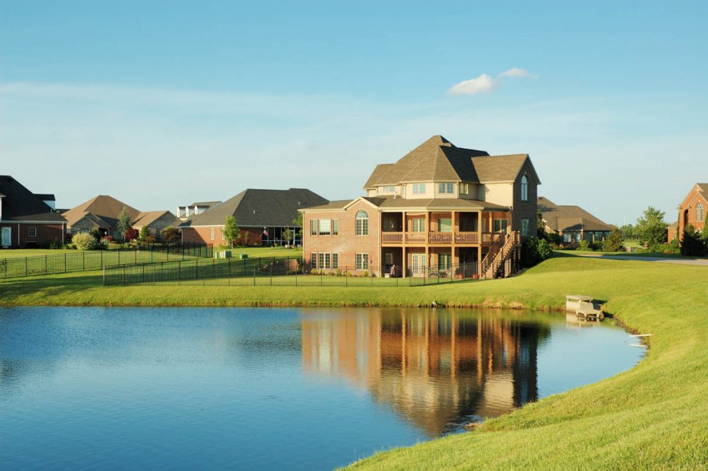 a three-story home surrounded by grass with a pond in the foreground