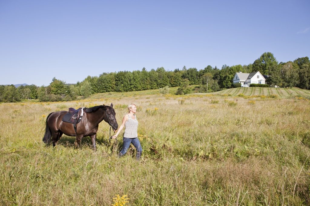 Woman leading horse through field with house in the distance